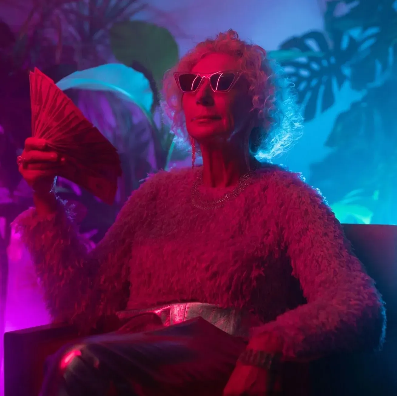 Woman in sunglasses holding a fan under pink and blue neon light