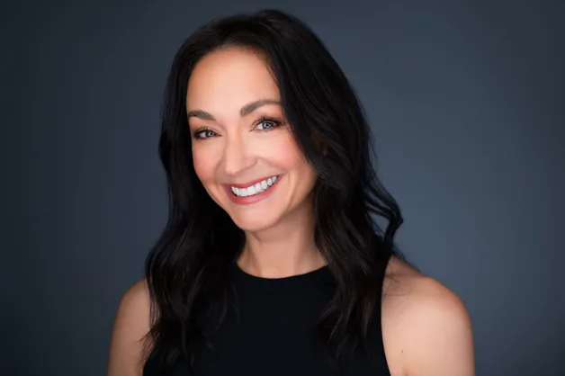 Smiling woman with long black hair wearing a black top, against a gray background.