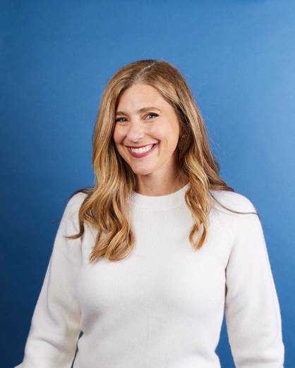 Close-up of a woman with long wavy light brown hair, smiling, wearing a white sweater, against a solid blue background.