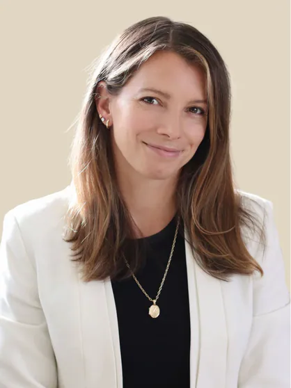 Young woman with light brown hair, wearing a white blazer, black top, gold necklace, and small gold earrings, smiling at the camera against a beige background.